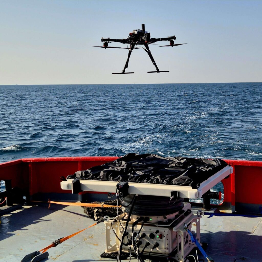 Large quadcopter drone hovering over an open sea, with a deck and scientific equipment in the foreground.