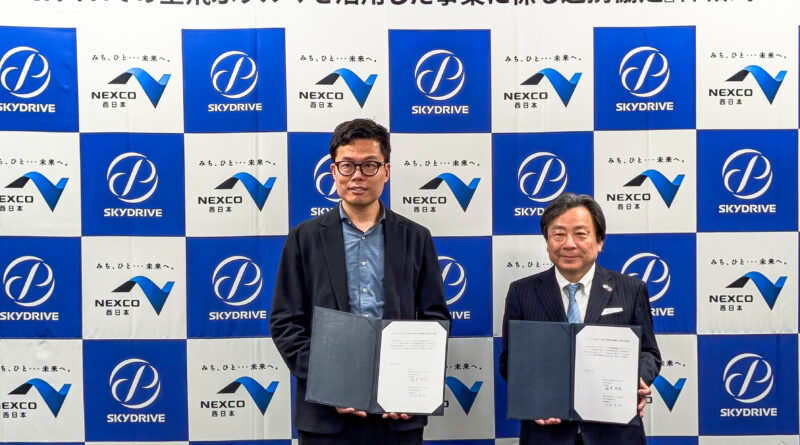 Two men in dark suits pose with open folders at a signing ceremony in front of a blue-and-white NEXCO SkyDrive backdrop.