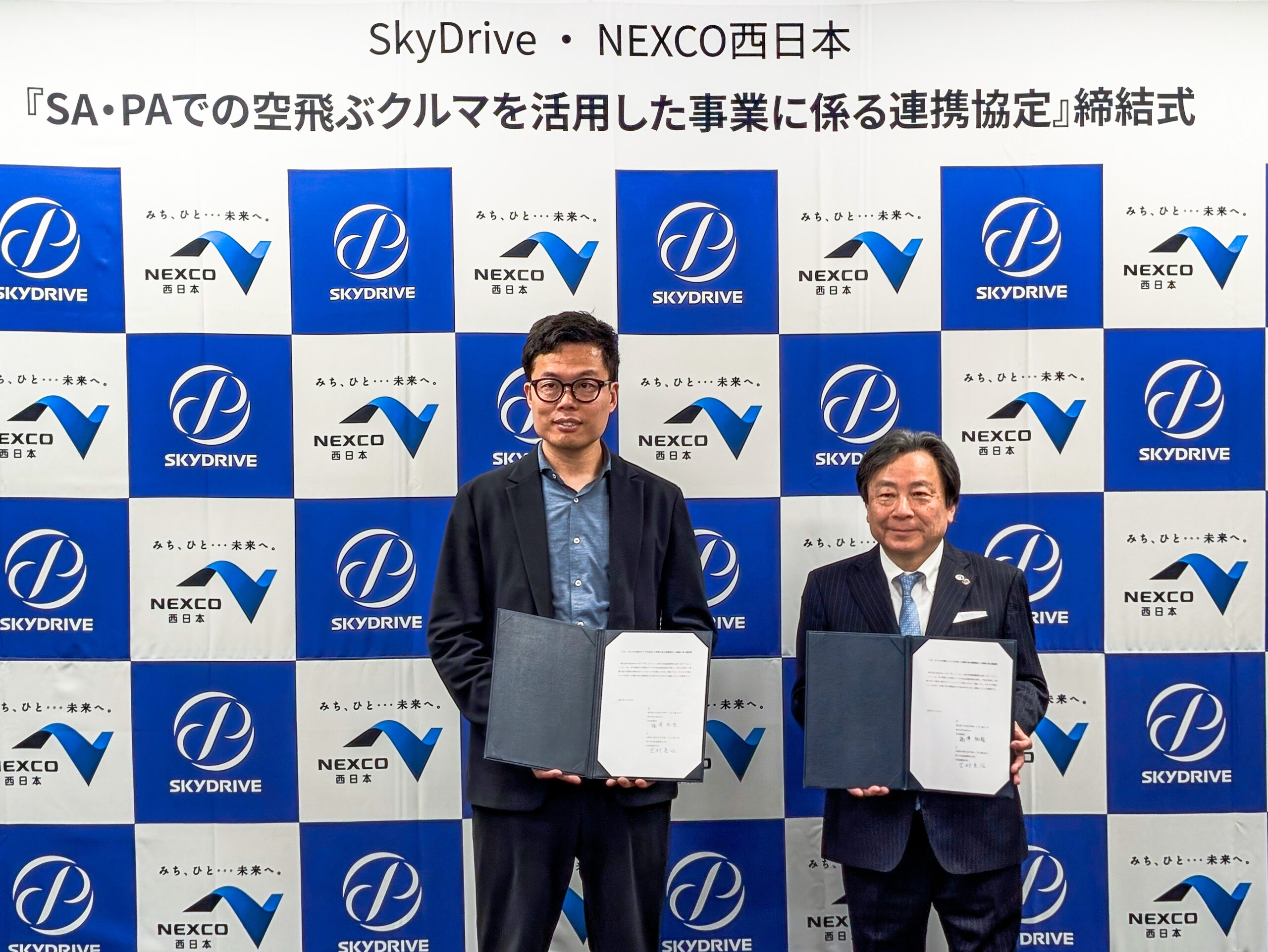 Two men in dark suits pose with open folders at a signing ceremony in front of a blue-and-white NEXCO SkyDrive backdrop.