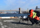 Person in a neon safety vest crouches beside a Skyports drone on a rooftop, with the Brooklyn Bridge and NYC skyline in the background.
