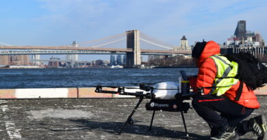 Person in a neon safety vest crouches beside a Skyports drone on a rooftop, with the Brooklyn Bridge and NYC skyline in the background.