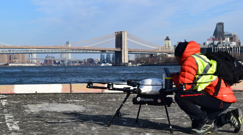 Person in a neon safety vest crouches beside a Skyports drone on a rooftop, with the Brooklyn Bridge and NYC skyline in the background.