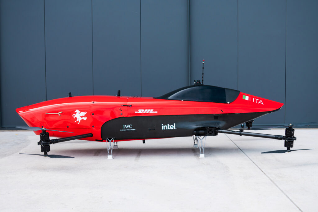 Red race hovercraft on landing gear, showing sponsor logos (IWC, Intel, DHL) and Italian flag against a dark gray wall.