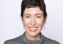 Smiling woman with short dark hair, visible green eyes, wearing a gray turtleneck sweater.
