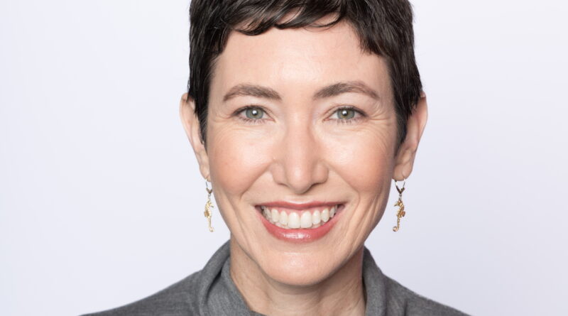 Smiling woman with short dark hair, visible green eyes, wearing a gray turtleneck sweater.