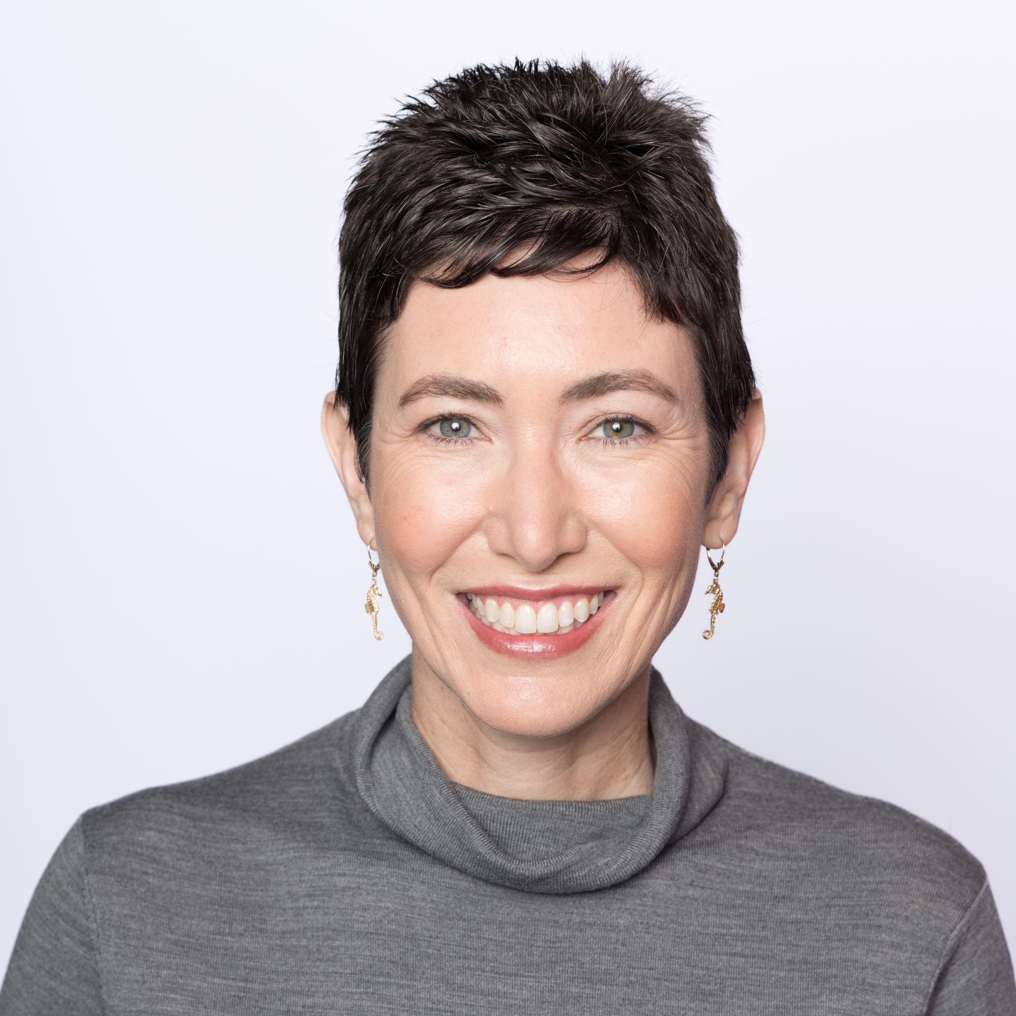 Smiling woman with short dark hair, visible green eyes, wearing a gray turtleneck sweater.