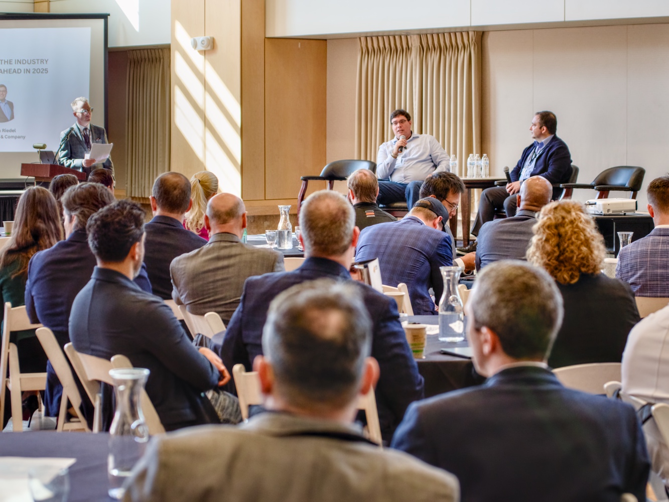 Conference panel discussion in a large room: a presenter at a podium on the left and two panelists seated with microphones on the right, facing an audience.