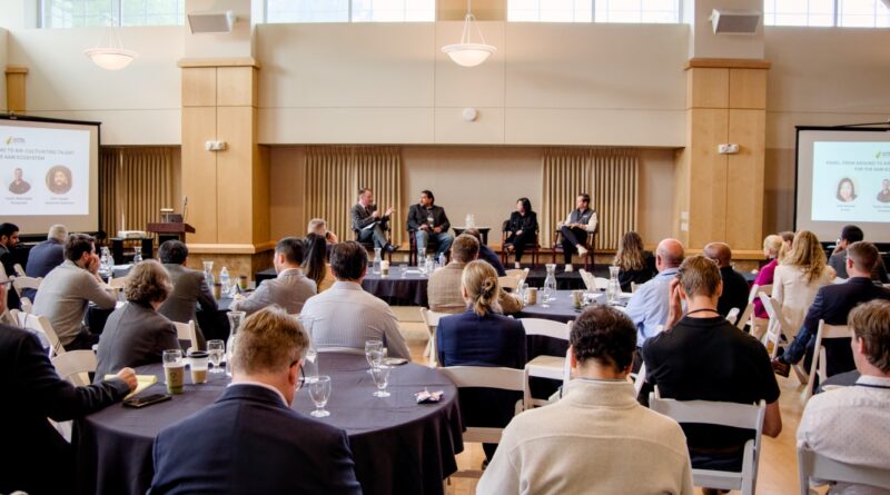 Panel discussion on a stage with four speakers, audience seated at round tables in a conference room.