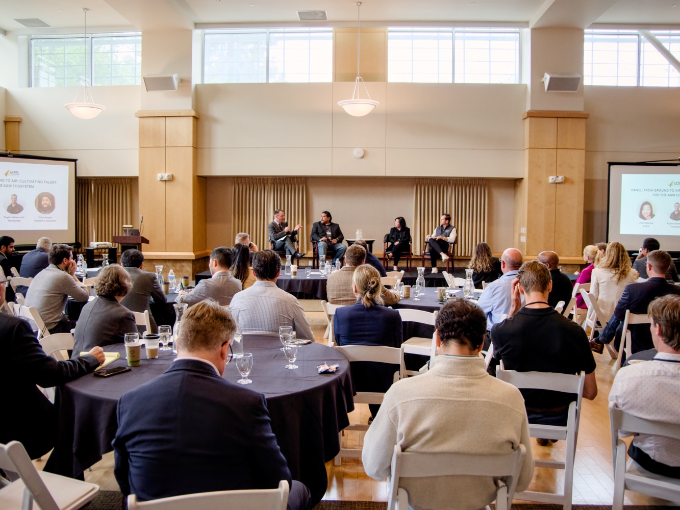 Panel discussion on a stage with four speakers, audience seated at round tables in a conference room.