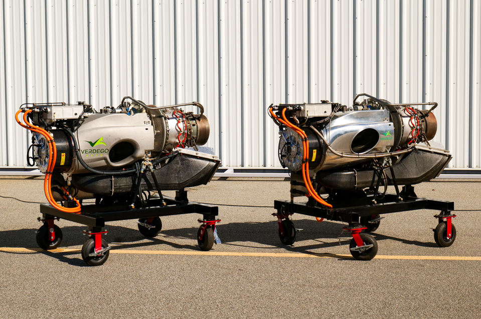 Two small jet engines on mobile stands side by side in a hangar, with orange hoses and a corrugated metal wall behind them (VERDEGO branding).