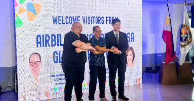 Three men stand on a stage, shaking hands in front of a large welcome banner; flags on the right and portraits on the backdrop hint at a formal event.