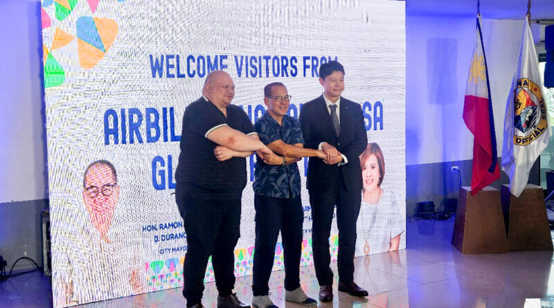 Three men stand on a stage, shaking hands in front of a large welcome banner; flags on the right and portraits on the backdrop hint at a formal event.