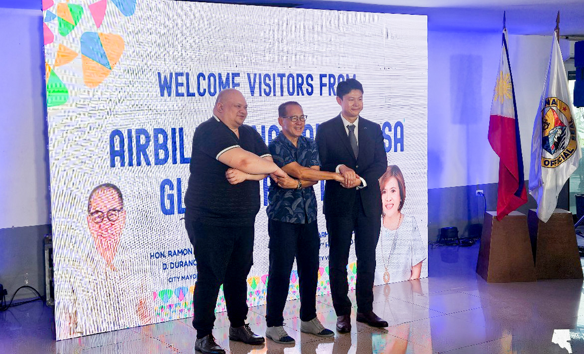 Three men stand on a stage, shaking hands in front of a large welcome banner; flags on the right and portraits on the backdrop hint at a formal event.