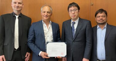 Four men in suits pose for a formal photo; the second man from the left holds a framed certificate as they stand against a wooden panel wall.
