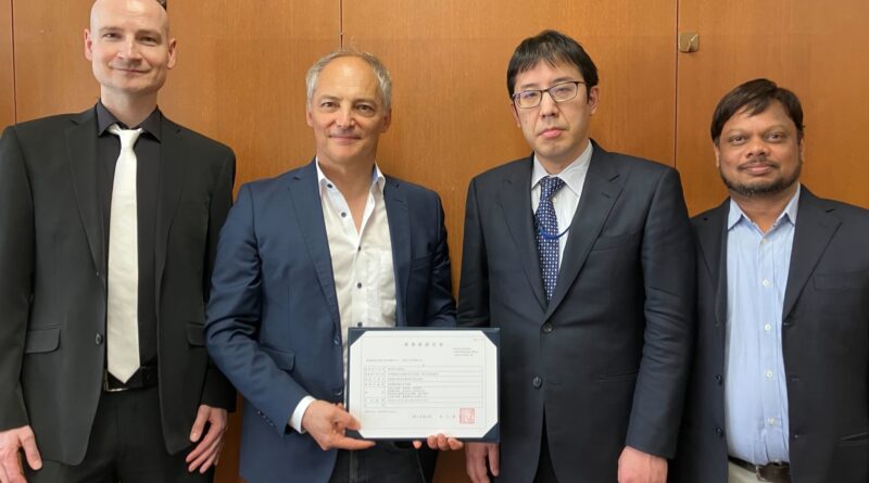 Four men in suits pose for a formal photo; the second man from the left holds a framed certificate as they stand against a wooden panel wall.