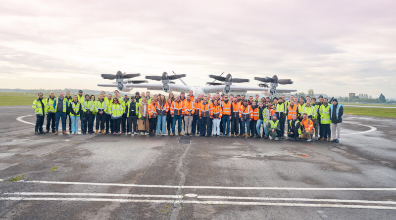Large group of airport workers in high-visibility vests posing on the tarmac with a propeller aircraft in the background, under a cloudy sky.
