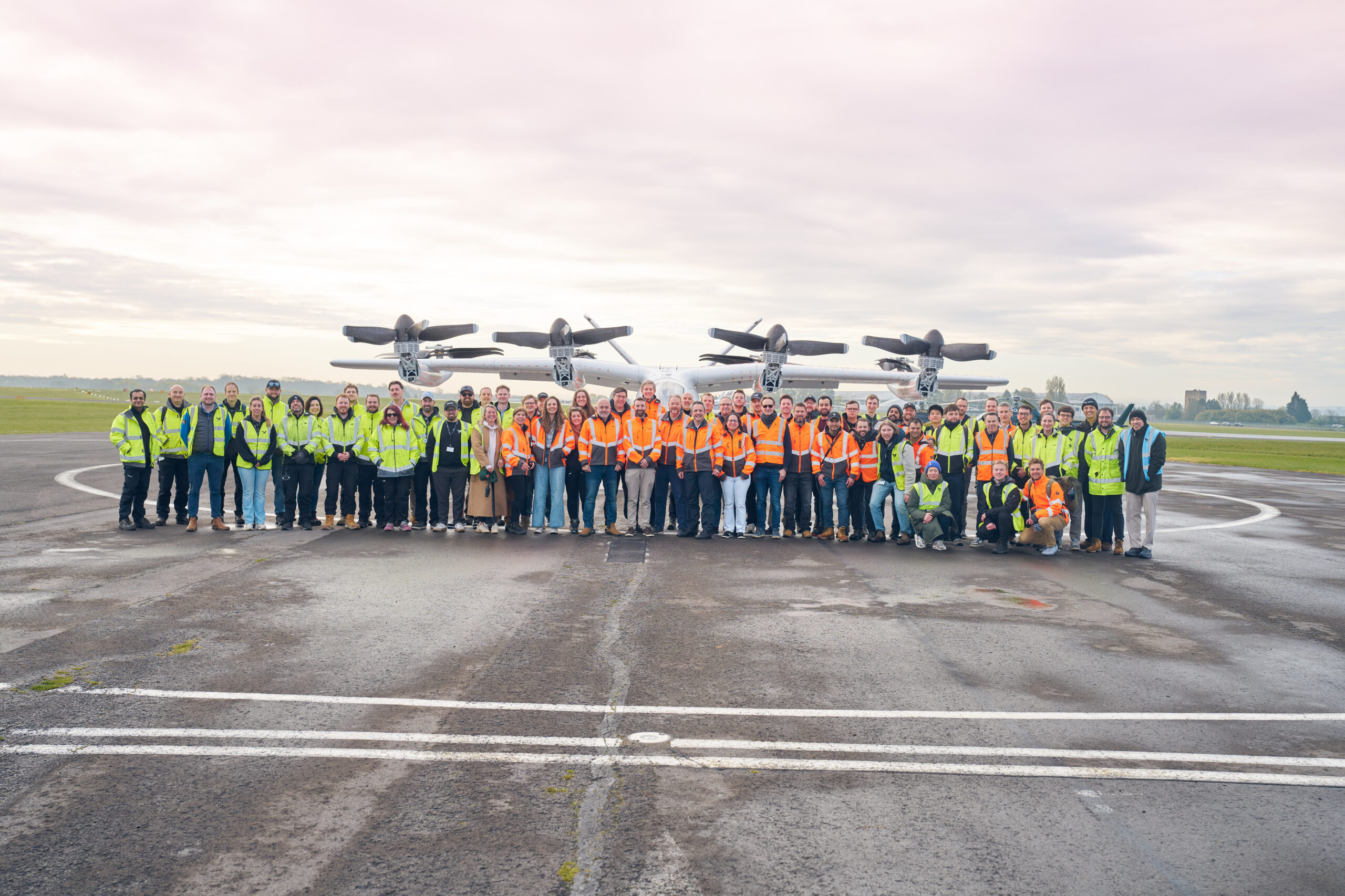 Large group of airport workers in high-visibility vests posing on the tarmac with a propeller aircraft in the background, under a cloudy sky.