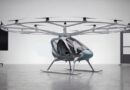 Futuristic Volocopter prototype with eighteen rotors above a teal-gray cabin in a showroom or gallery setting.