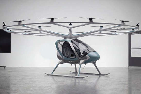 Futuristic Volocopter prototype with eighteen rotors above a teal-gray cabin in a showroom or gallery setting.