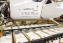 White aircraft fuselage with the 'smartflyer' logo and motto, on a production line with rows of battery packs or modules beneath it.