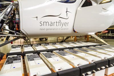 White aircraft fuselage with the 'smartflyer' logo and motto, on a production line with rows of battery packs or modules beneath it.