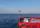Red boat deck with equipment; a drone flies over calm blue sea and distant ships on the horizon.