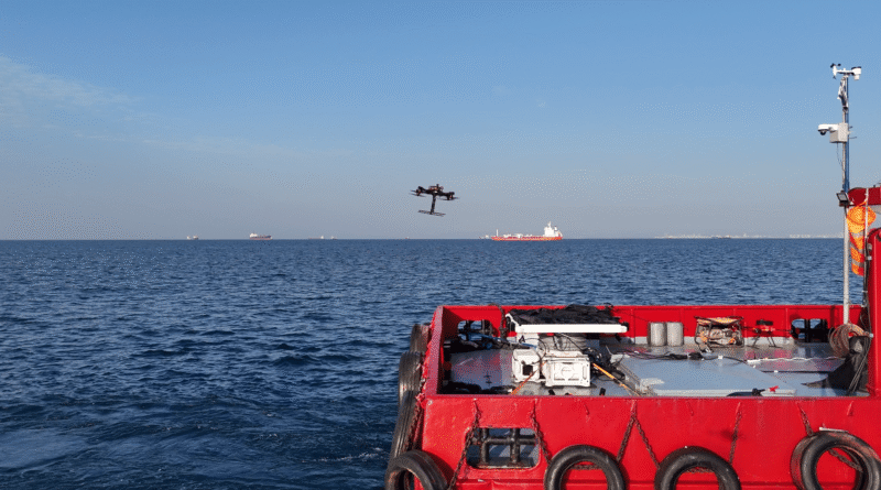 Red boat deck with equipment; a drone flies over calm blue sea and distant ships on the horizon.