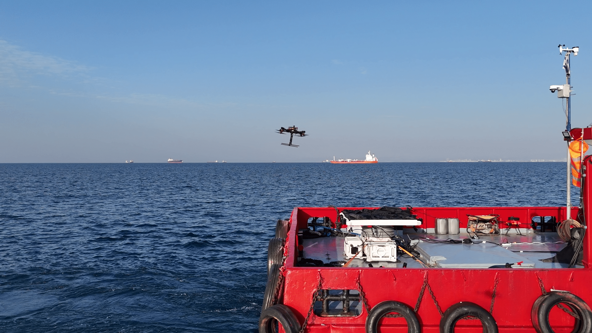 Red boat deck with equipment; a drone flies over calm blue sea and distant ships on the horizon.