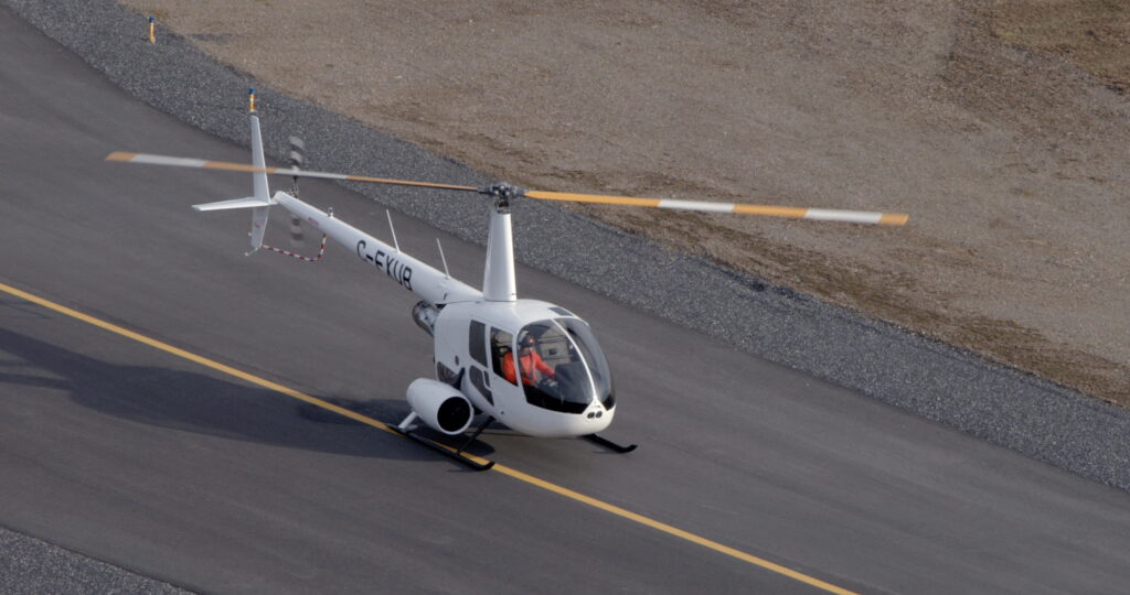 White helicopter on a paved road with a pilot in a red jacket visible in the cockpit, hillside in the background.