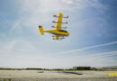 Yellow twin-engine aircraft flying low over an airfield with hangars in the distance under a blue sky, "wish" written in yellow at the bottom right.