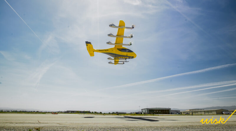 Yellow twin-engine aircraft flying low over an airfield with hangars in the distance under a blue sky, "wish" written in yellow at the bottom right.