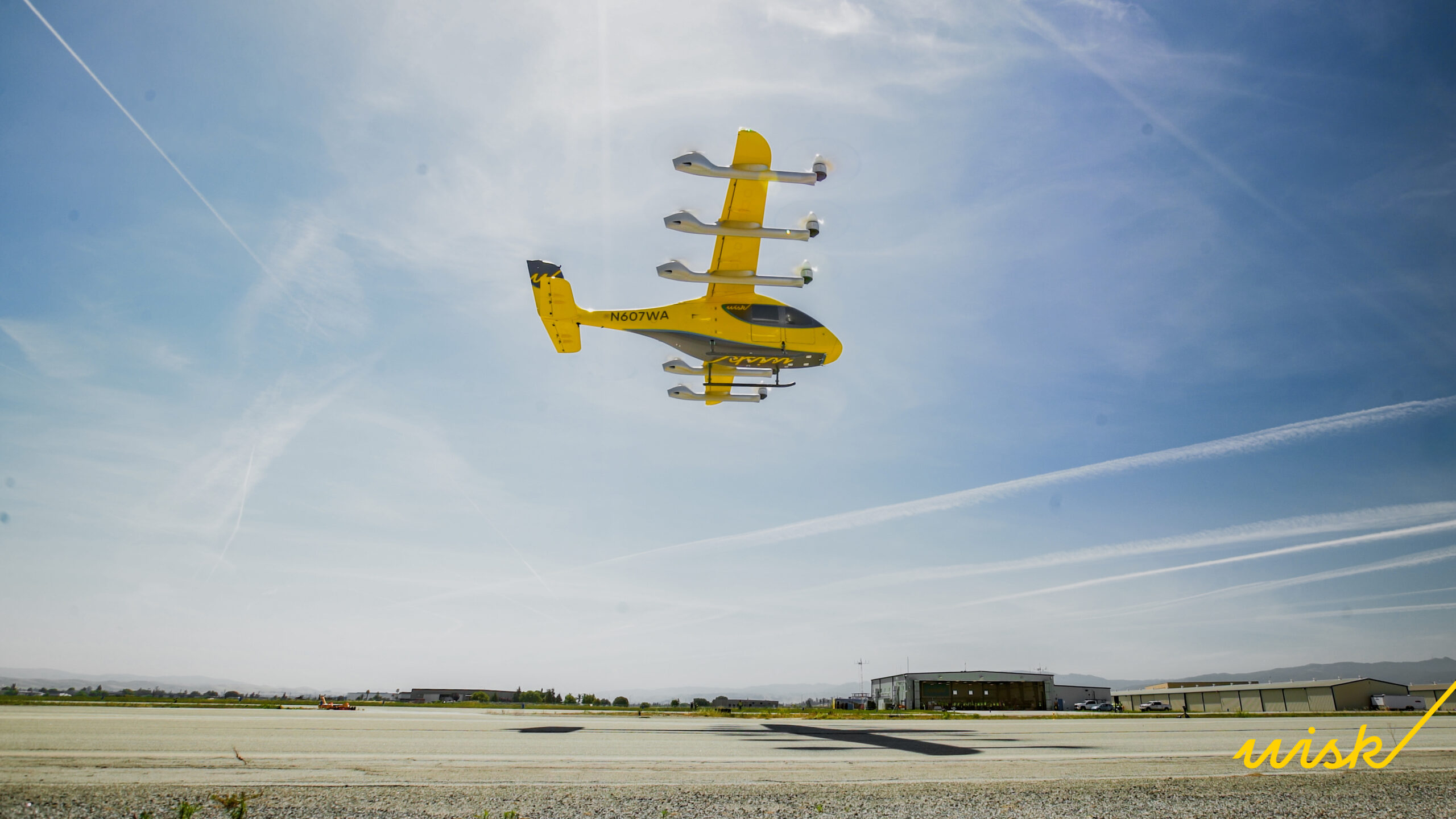 Yellow twin-engine aircraft flying low over an airfield with hangars in the distance under a blue sky, "wish" written in yellow at the bottom right.