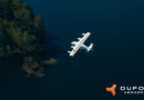 White twin-engine airplane flying over dark blue water, forested shoreline on the left, DUFOUR Aerospace logo bottom right.