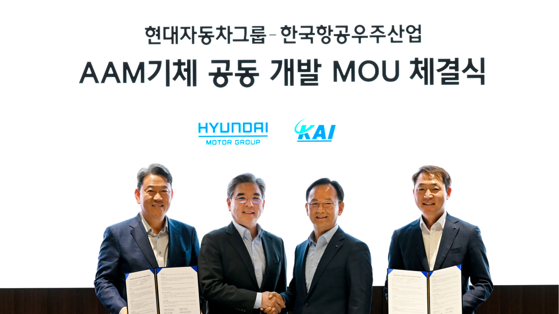 Four executives in suits celebrate a MOA signing between Hyundai Motor Group and KAI, holding signed documents under a Korean banner and logos above.
