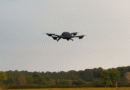 Quadcopter drone with green and red navigation lights flying low over a grassy field and treeline beneath a cloudy sky.