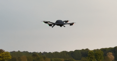 Quadcopter drone with green and red navigation lights flying low over a grassy field and treeline beneath a cloudy sky.