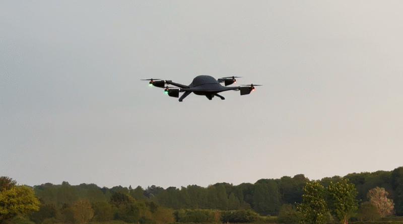 Quadcopter drone with green and red navigation lights flying low over a grassy field and treeline beneath a cloudy sky.