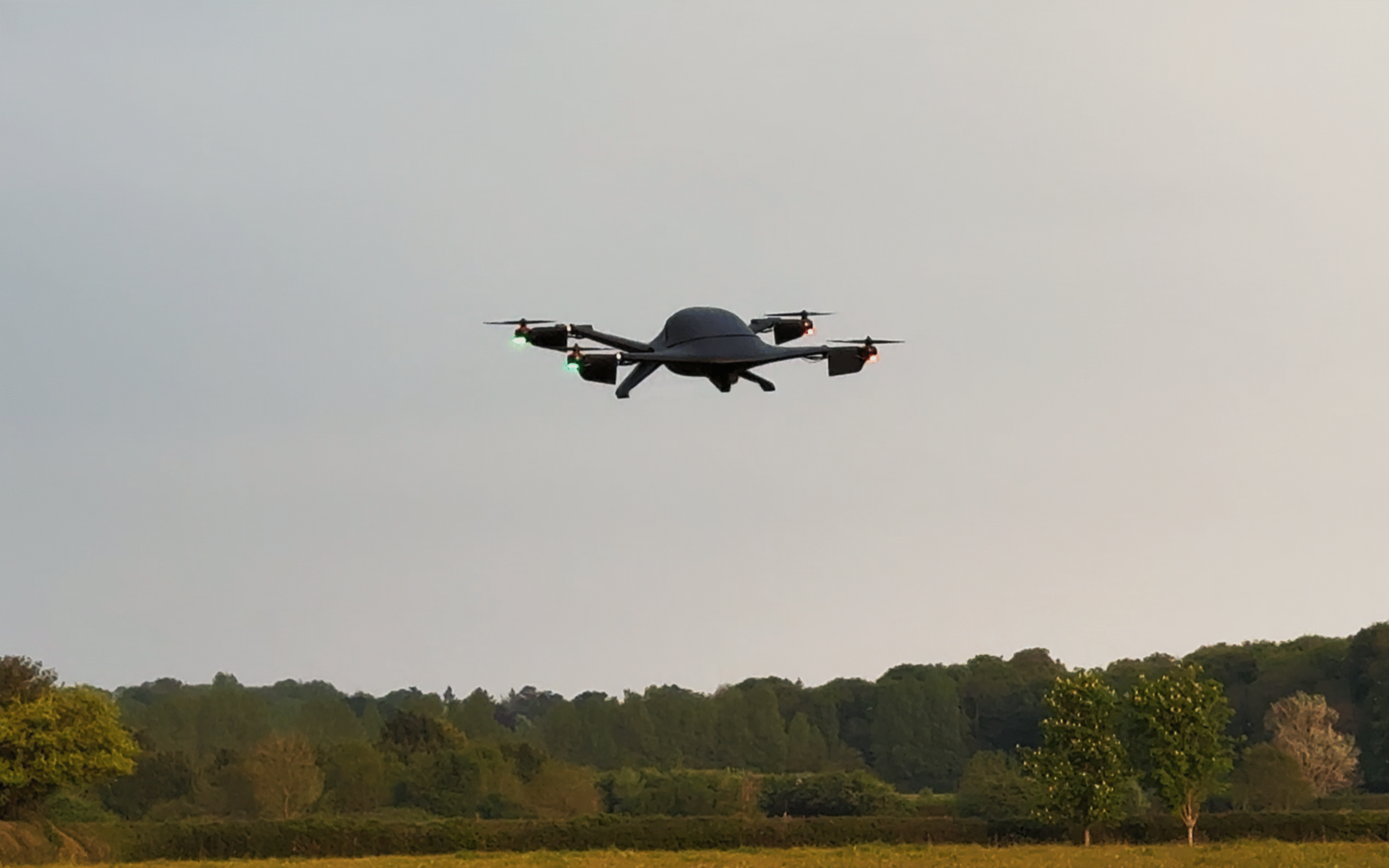 Quadcopter drone with green and red navigation lights flying low over a grassy field and treeline beneath a cloudy sky.