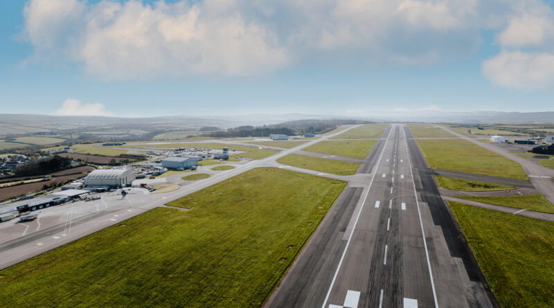 Cornwall Airport Newquay - eVTOL Insights Aerial view of a rural airport showing a long runway with multiple taxiways, hangars, and surrounding green fields under a blue sky.