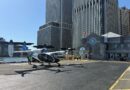 Twin-engine helicopter parked on a tarmac next to water and tall office buildings; people in safety vests nearby at a city heliport, blade aircraft logo visible on fuselage