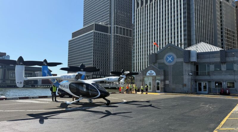 Twin-engine helicopter parked on a tarmac next to water and tall office buildings; people in safety vests nearby at a city heliport, blade aircraft logo visible on fuselage