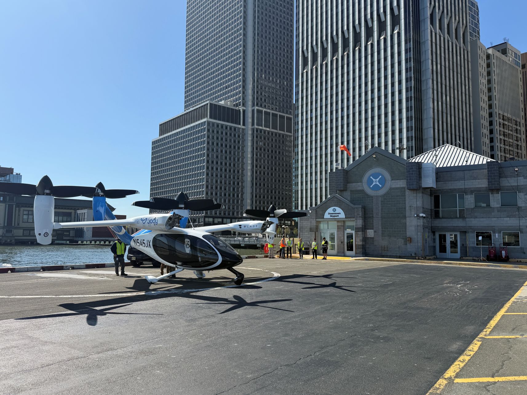 Twin-engine helicopter parked on a tarmac next to water and tall office buildings; people in safety vests nearby at a city heliport, blade aircraft logo visible on fuselage