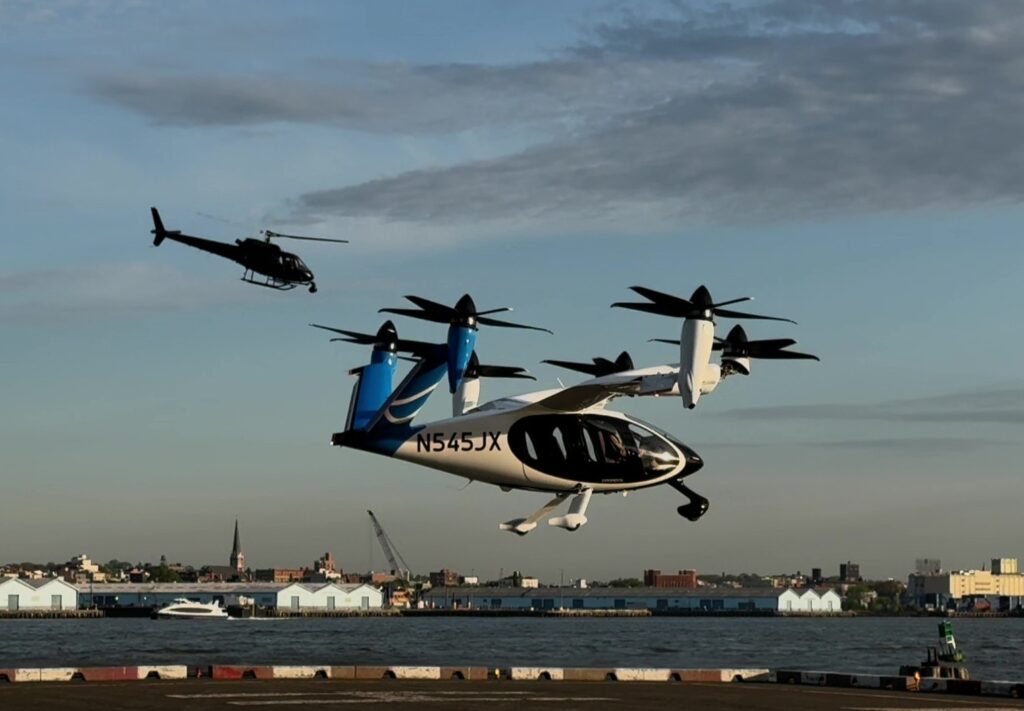 White and blue tiltrotor aircraft (V-22 Osprey) lifting off over a harbor, with a small helicopter in the sky behind it.