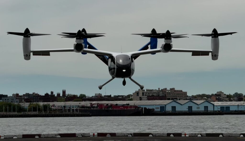 Front view of a quad-rotor VTOL aircraft hovering low over water with a city waterfront in the background.