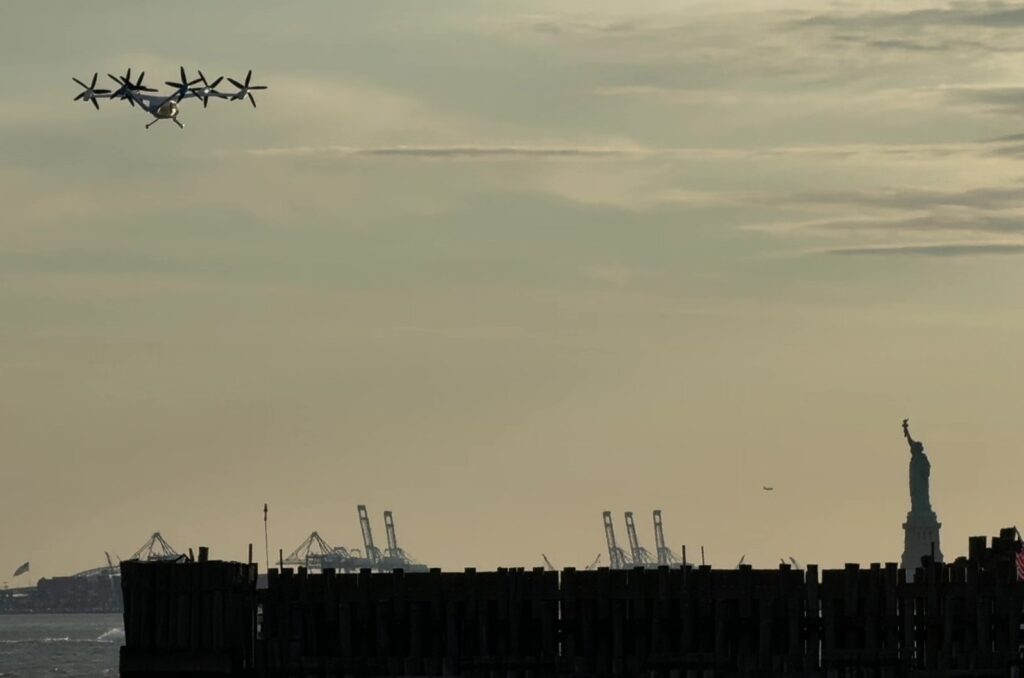 Silhouetted six‑propeller aircraft flying low over a harbor at sunset, with the Statue of Liberty visible in the distance and a wooden breakwater in the foreground.