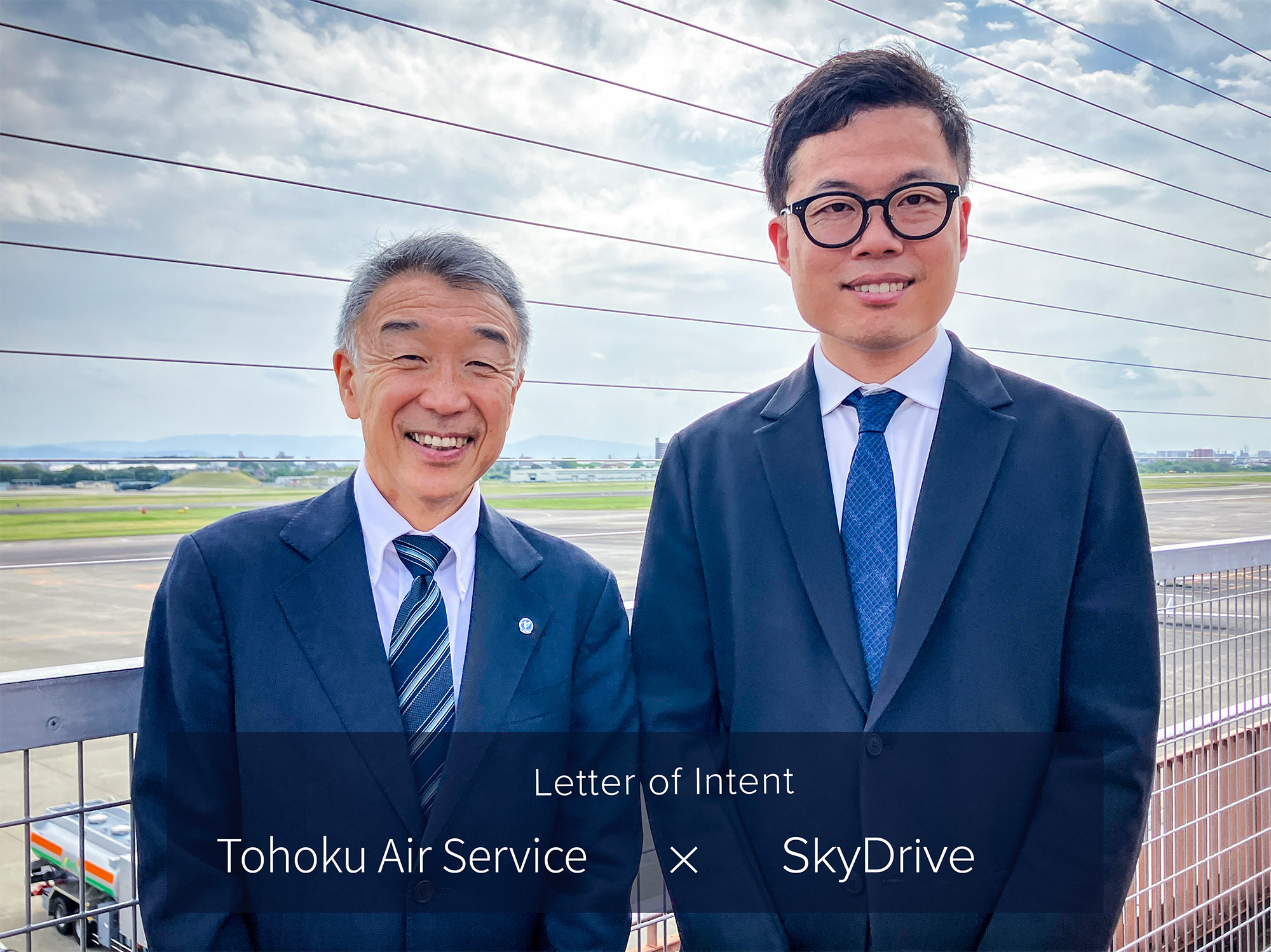 Two men in suits smiling on a rooftop at an airfield, announcing a partnership between Tohoku Air Service and SkyDrive.