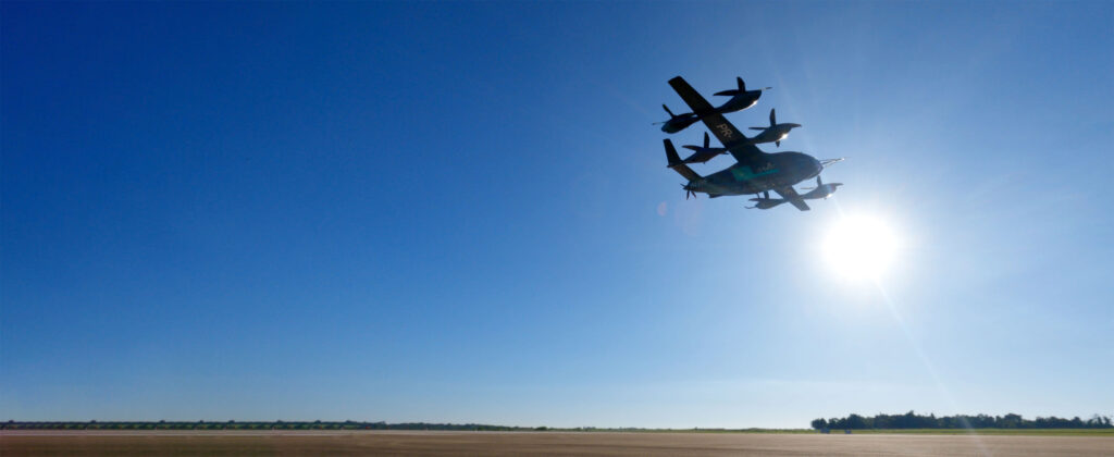 Tiltrotor aircraft (Osprey) flying low over a runway with a bright sun in a clear blue sky, horizon visible below.