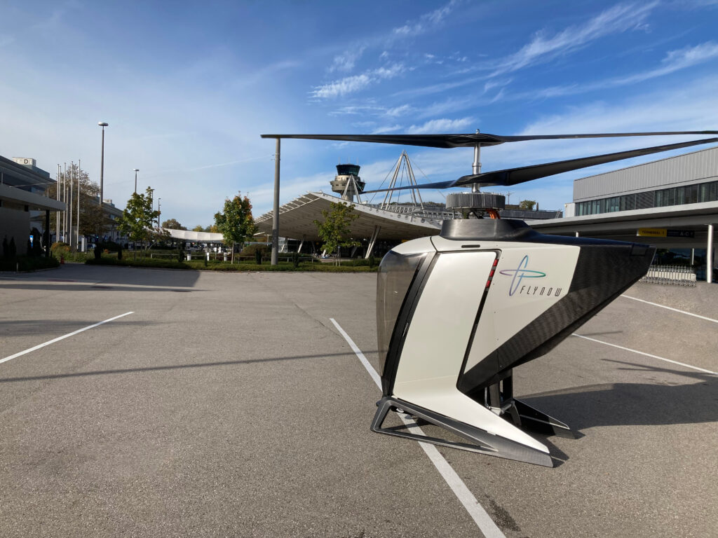 White helicopter parked on an airport tarmac with a distant control tower and modern buildings under a blue sky.