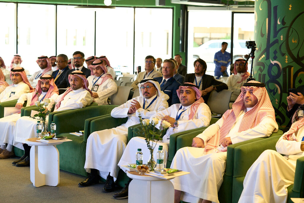 Audience of men in traditional Middle Eastern dress seated on green sofas at a conference, with tables holding water bottles and flowers in front of them.