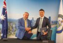 Two men in business suits shake hands in front of a city skyline backdrop, each holding a folder or document.